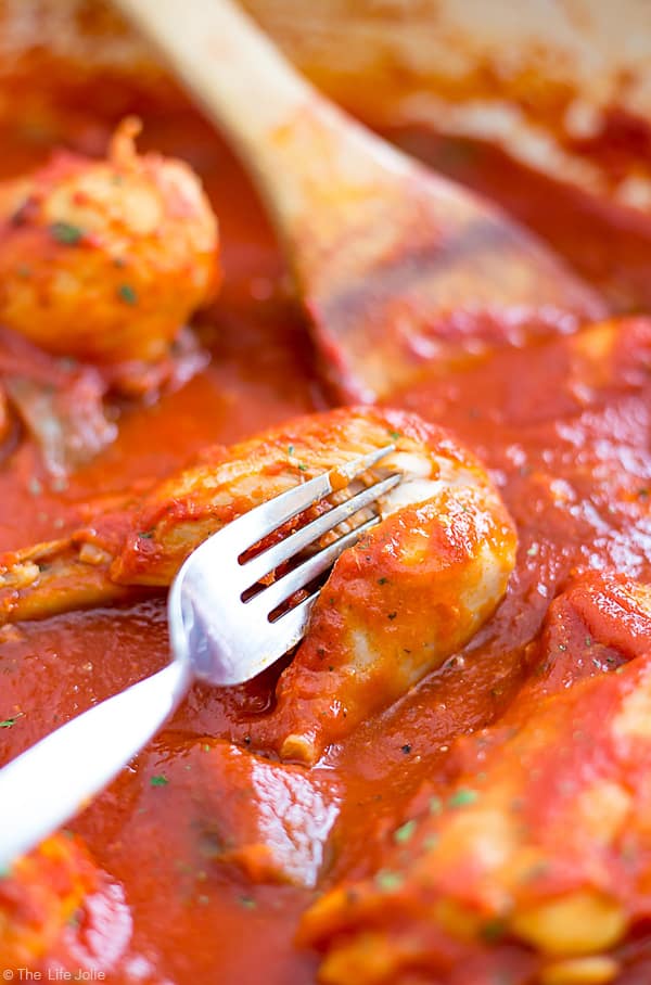 Pieces of chicken in cacciatore sauce in a pan with a wooden spoon in the background and a fork poking into a piece of the chicken showing how tender it is.