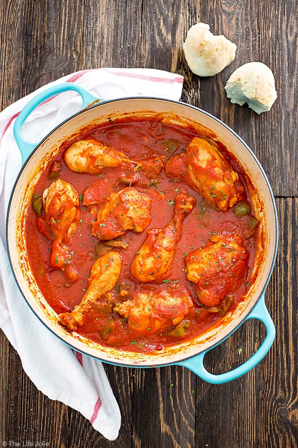 An overhead image of a pan of chicken cacciatore with a wooden spoon in it on top of a white towel with a couple red stripes and some garlic bulbs next to it.