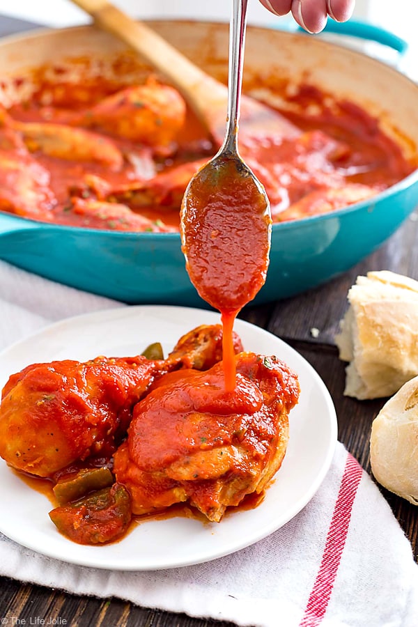 A spoon pouring extra cacciatore sauce on top of chicken pieces on a plate with a pan of chicken behind it and bread on the side.