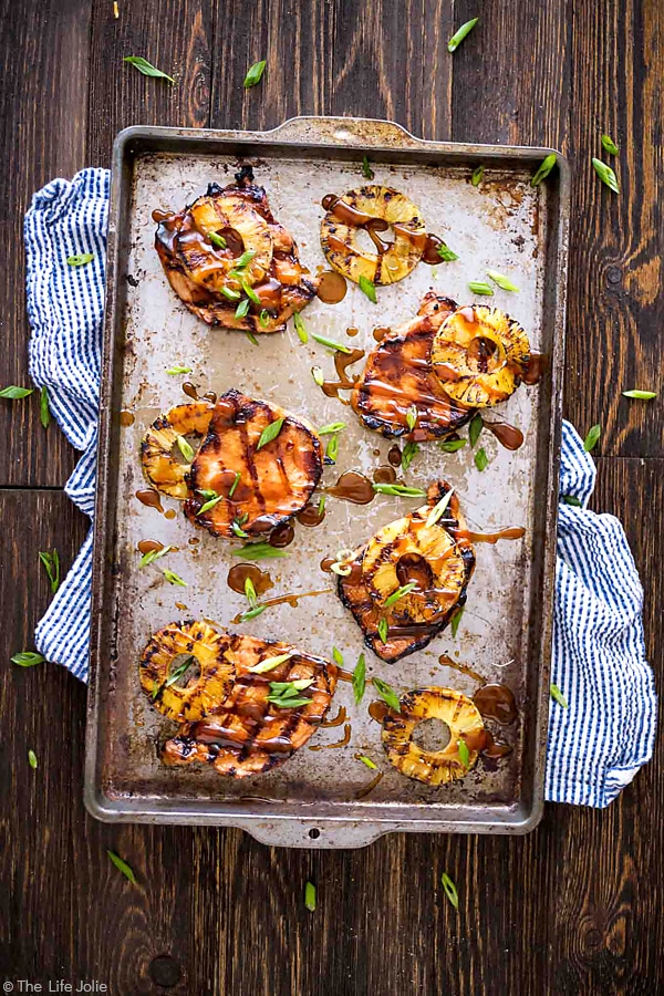 An overhead image of a pan of grilled pork chops and pineapple on a blue and white striped towel with a wood background.