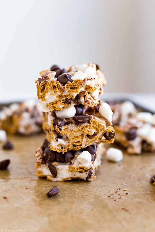 A straight-on image of 3 s'mores bars stacked on top of each other with other bars behind them and chocolate chips and marshmallows.