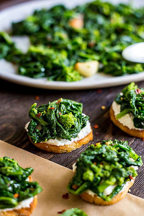 An image of Broccoli Rabe and Ricotta Crostini with a blurry crostini in the foreground and the blurry plate of broccoli rabe in the background.