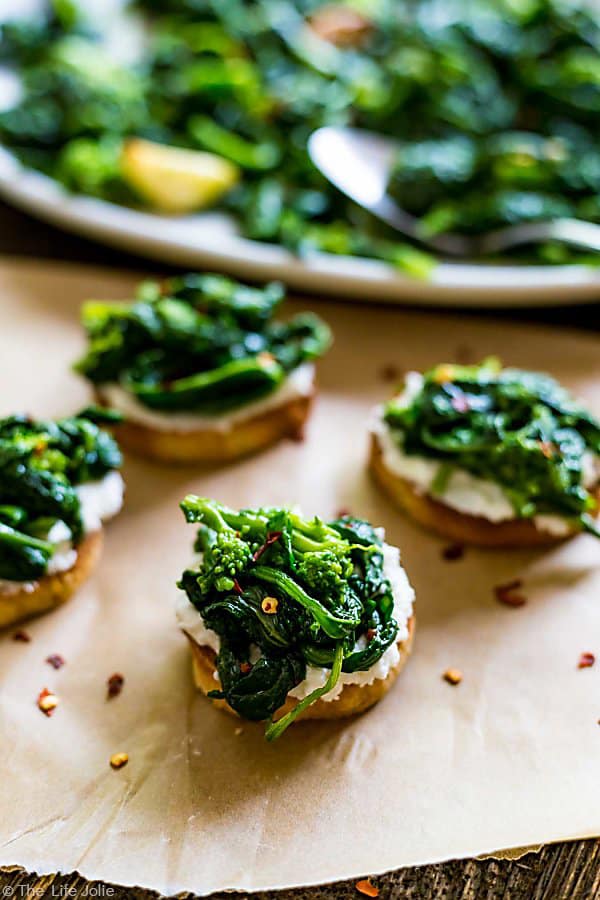 A couple Broccoli Rabe and Ricotta Crostini's on a piece of parchment paper with crushed red pepper flakes surrounding it and a plate of broccoli rabe in the background.