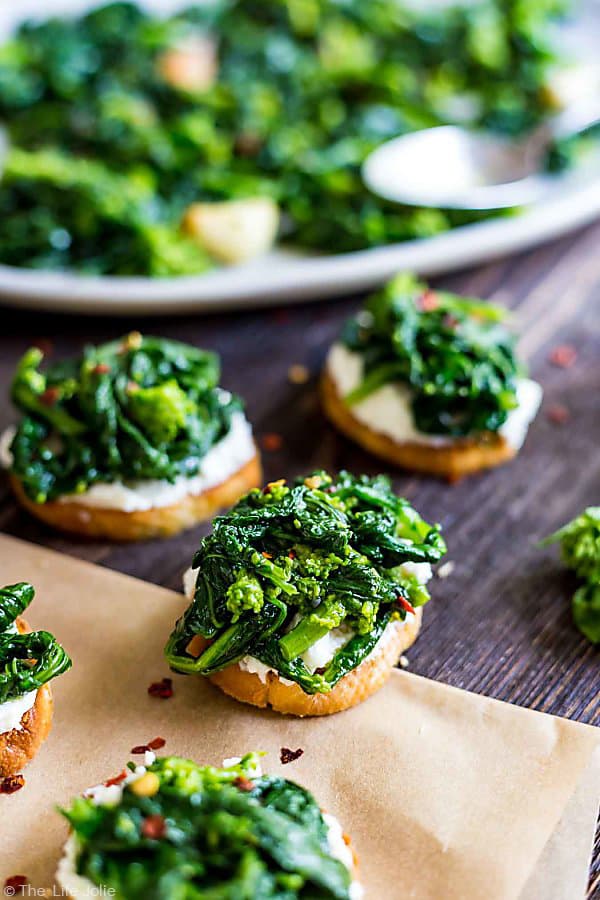 A Broccoli Rabe and Ricotta Crostini front and center with two out of focus crostinis in the background and a plate of broccoli rabe.