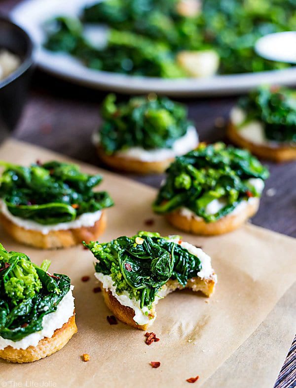 A piece of Broccoli Rabe and Ricotta Crostini with a bite taken out of it in the front and other crostinis in the background and out of focus.