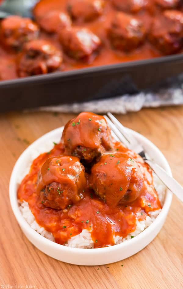 A plate of three Classic Porcupine Meatballs over white rice with a pan of other meatballs out of focus behind it.