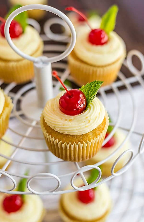 A close up of a Mai Tai Cupcake on a cupcake stand with a cherry on top.