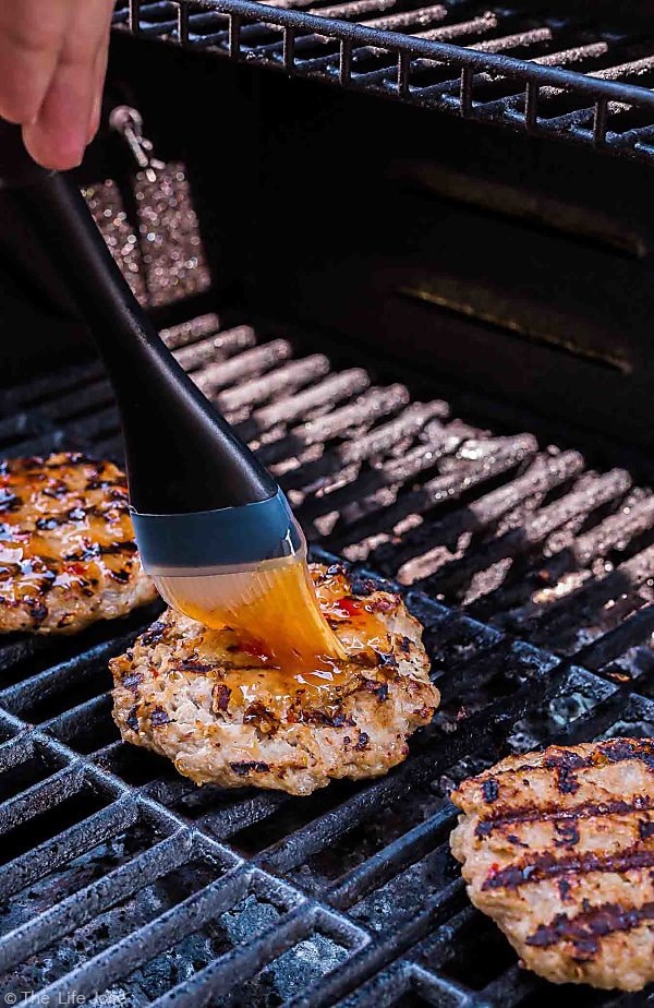 A close up shot of a brush coming from the top left corner brushing Frank's RedHot Sweet Chili sauce onto the Sweet Chili Turkey Burgers on thew grill.