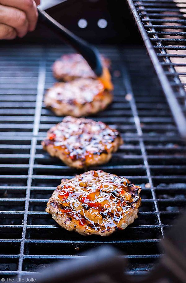 A shot of the four Sweet Chili Turkey Burgers on the grill with the front burger in focus and a brush basting the third burger in the background out of focus.