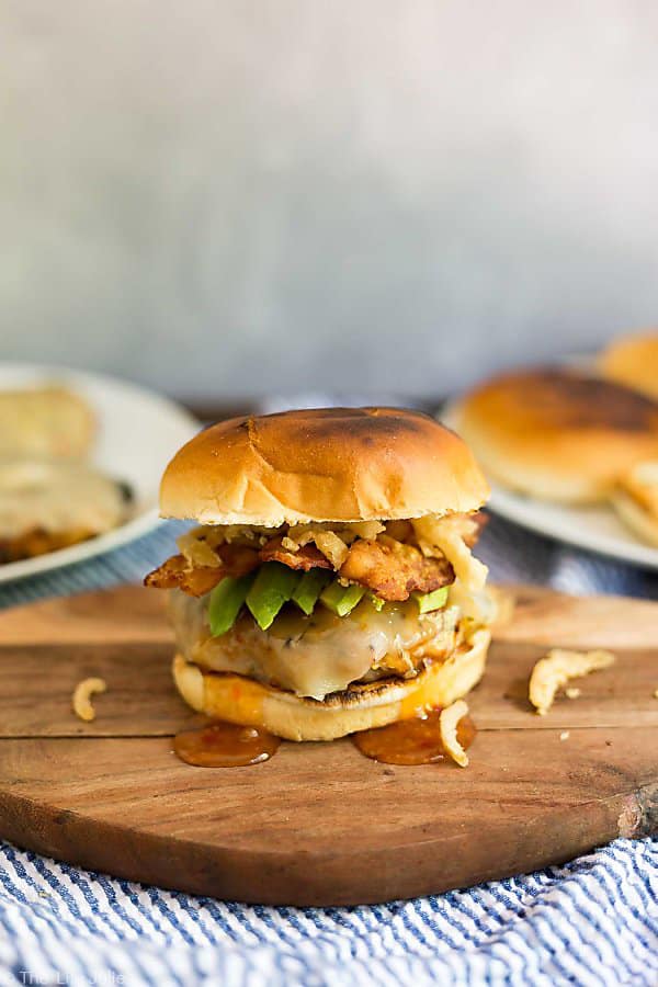 A straight on shot of a Sweet Chili Turkey Burger on a cutting board with a plate of grilled buns and a plate of burgers in the background out of focus.
