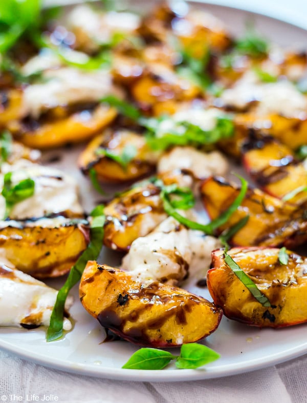 A close up image of a peach in the Grilled Peaches and Burrata Salad with balsamic drizzled on it and fresh basil leaves with the rest of the salad behind it and out of focus.