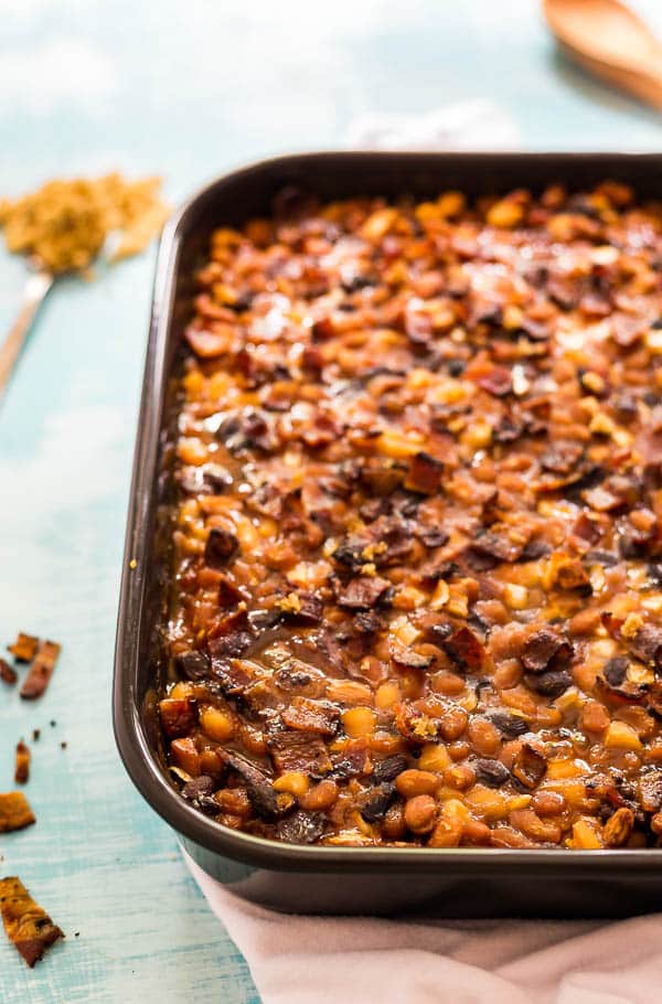 A shot of the left side of a pan of Most Delicious Baked Beanswith a spoon of brown sugar and some bacon crumbled on the side of it.