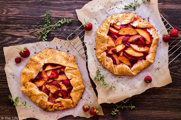 An overhead image of two Nectarine and Strawberry Galette's with Thymeon parchment paper with a wood background and fresh thyme and strawberries surrounding it,