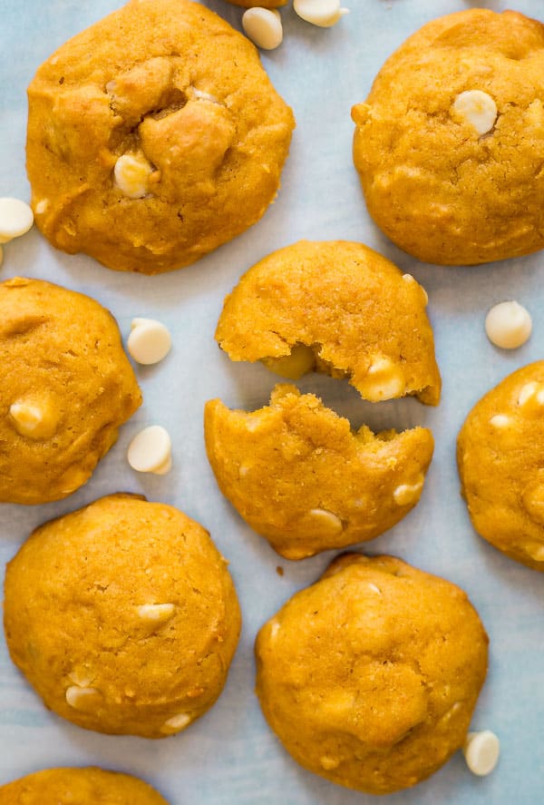 An overhead shot of White Chocolate Macadamia Nut Pumpkin Cookieswith white chocolate chips around them and one cookie broken into two pieces in the middle.