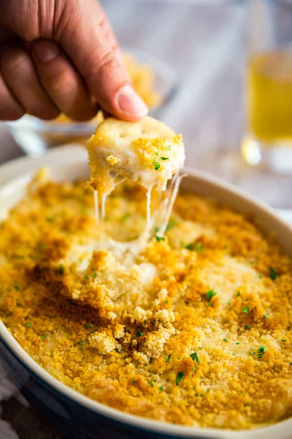 A hand pulling up a cracker full of Hot Chicken Swiss Dip from a pan of the dip with stringy cheese trailing.