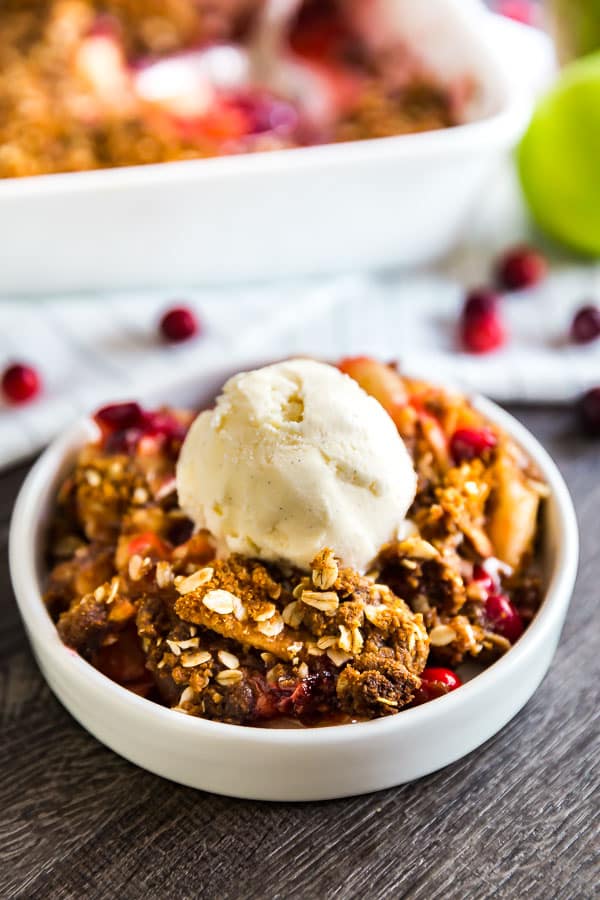 A round white plate with Cranberry Apple Crisp Recipe with Oatmeal Ginger Snap Topping topped with vanilla ice cream and a pan of it in the background.