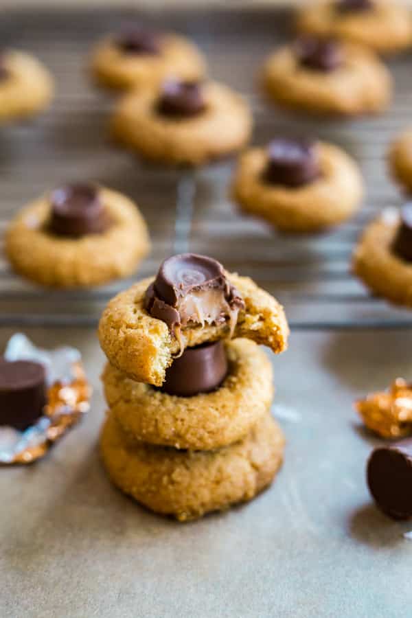 A stack of Salted Caramel Blossoms Cookies with a bite taken out of the top one and a cooling rack with more of them on it in the background.